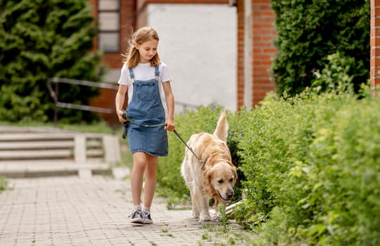 Preteen girl wearing jeans dress with golden retriever dog walking outdoors in summertime. Pretty kid petting fluffy doggy pet in city