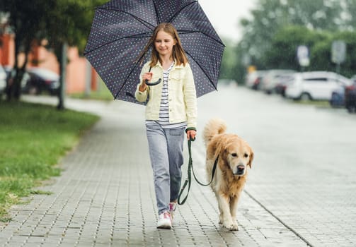 Preteen girl with golden retriever dog in rainy day walking outdoors in city park. Cute kid child with labrador pet doggy outside