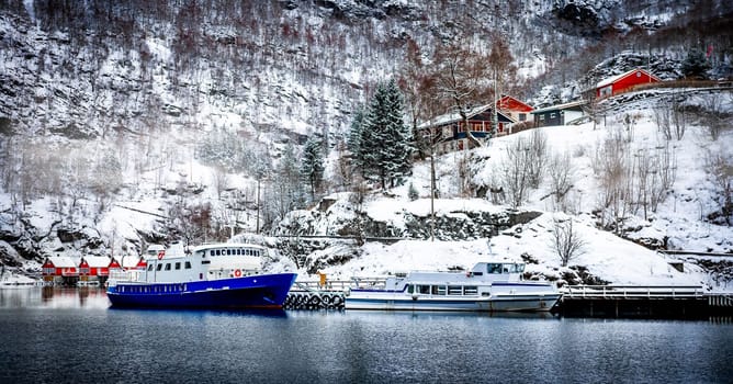 Charming Norway lake view with boats and wooden red houses on snowy hill in winter. Scenery scandinavian landscape with sea and beautiful reflection in Europe