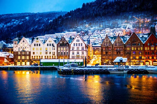 Winter Bergen city with famous Bryggen merchandise wooden houses and lights in snow season. Panorama of historical harbor buildings at Christmas time with magical reflection in sea