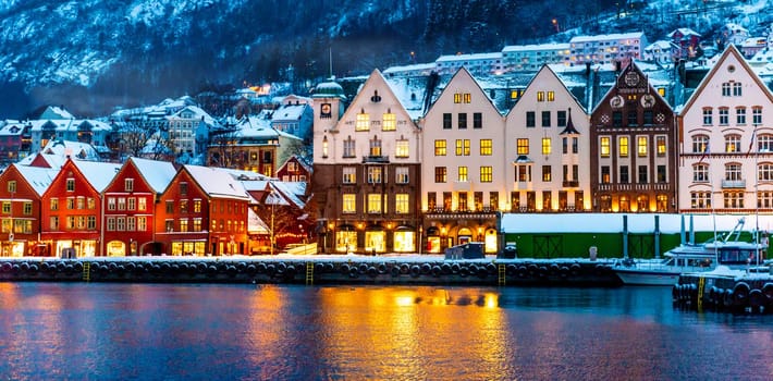 Winter Bergen city with famous Bryggen merchandise wooden houses and lights in snow season. Panorama of historical harbor buildings at Christmas time with magical reflection in sea