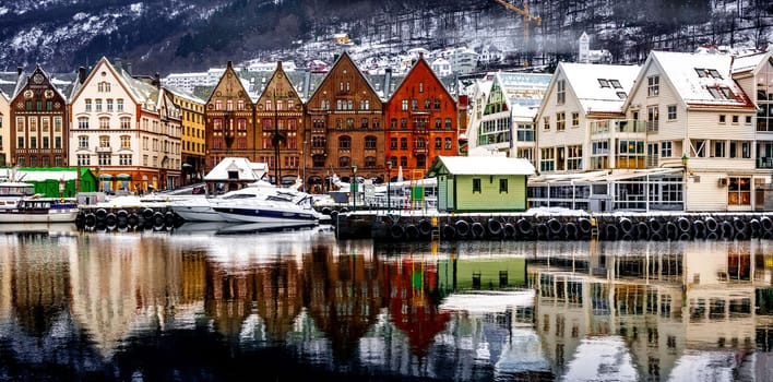 Winter Bergen city with famous Bryggen UNESCO merchandise wooden houses in snow season. Panorama of historical harbor buildings at Christmas time with magical reflection in sea