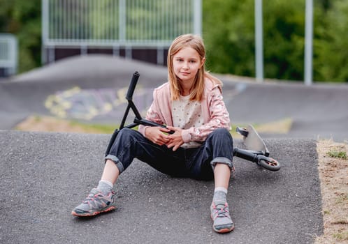 Preteen girl with scooter sitting in city park at spring time and looking at camera. Pretty child kid model posing with vehicle