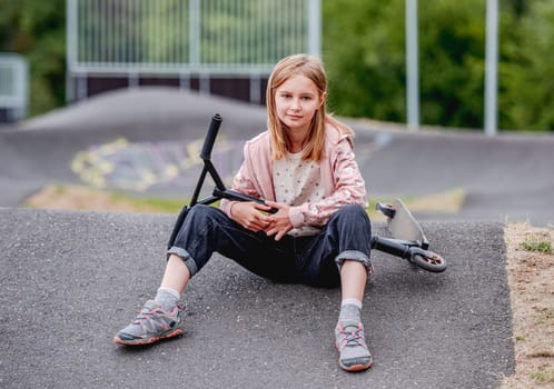 Preteen girl with scooter sitting in city park at spring time and looking at camera. Pretty child kid model posing with vehicle