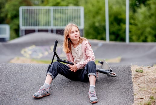 Preteen girl with scooter sitting in city park at spring time and looking at camera. Pretty child kid model posing with vehicle