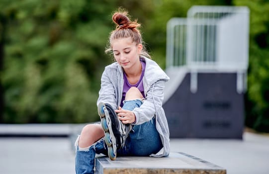 Cute girl roller skater sitting in city park and smiling. Pretty female teenager posing during rollerskating