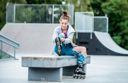 Cute girl roller skater sitting in city park and smiling. Pretty female teenager posing during rollerskating