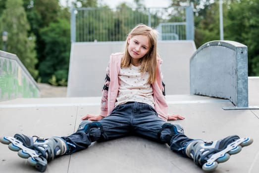 Cute girl roller skater sitting in city park on ground. Pretty female preteen kid child posing during rollerskating