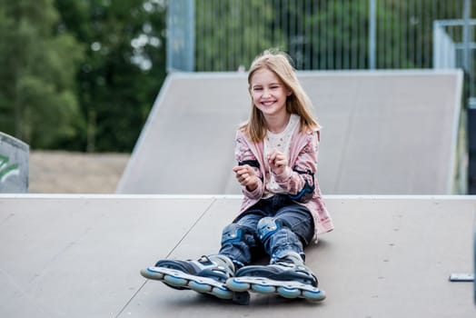 Cute girl roller skater sitting in city park and smiling. Pretty female preteen kid child posing during rollerskating
