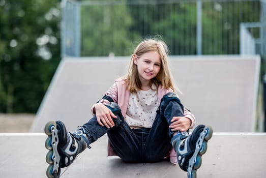 Cute girl roller skater sitting in city park on ground. Pretty female preteen kid child posing during rollerskating