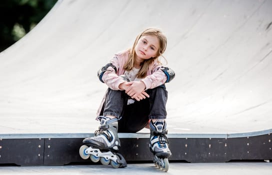 Cute girl roller skater sitting in city park and looking at camera Pretty female preteen kid child posing during rollerskating