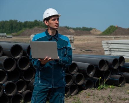 Caucasian male builder in a hard hat stands near the pipes and uses a laptop at a construction site