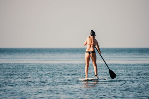Sea woman sup. Silhouette of happy middle aged woman in rainbow bikini, surfing on SUP board, confident paddling through water surface. Idyllic sunset. Active lifestyle at sea or river