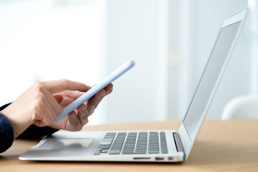 Side view of woman hands using mobile phone while working in the office with laptop. Blurred background. Texting concept.