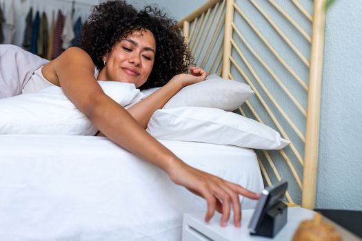 African American young woman waking up happy after good night sleep reaching hand to turn off alarm clock. Lifestyle concept.