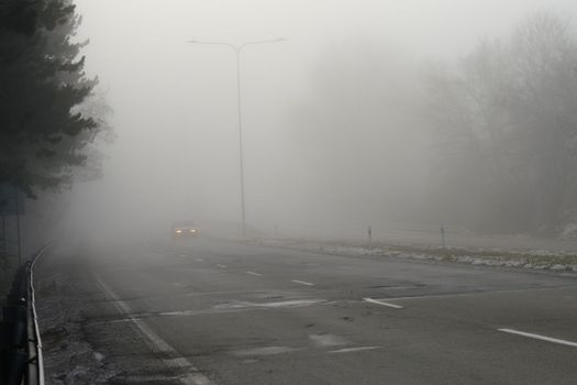 Car traveling on the foggy road with headlights or headlamps on. Low visibility - Dangerous driving of cars in winter in bad weather.