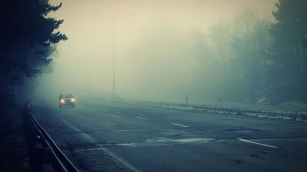 Car traveling on the foggy road with headlights or headlamps on. Low visibility - Dangerous driving of cars in winter in bad weather.