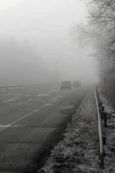 Car traveling on the foggy road with headlights or headlamps on. Low visibility - Dangerous driving of cars in winter in bad weather.