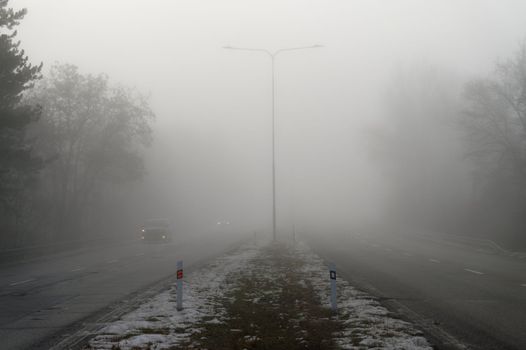 Car traveling on the foggy road with headlights or headlamps on. Low visibility - Dangerous driving of cars in winter in bad weather.