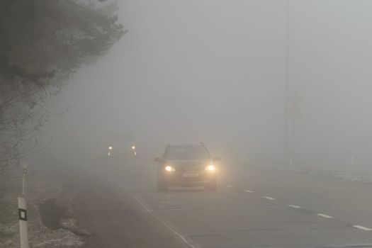 Car traveling on the foggy road with headlights or headlamps on. Low visibility - Dangerous driving of cars in winter in bad weather.