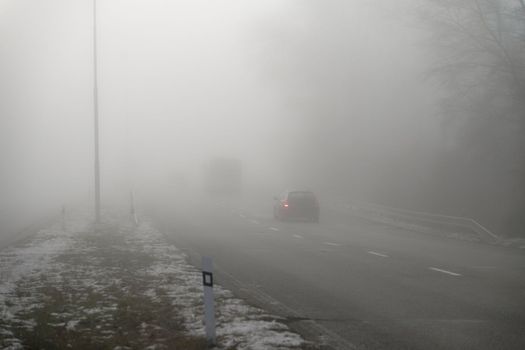 Car traveling on the foggy road with headlights or headlamps on. Low visibility - Dangerous driving of cars in winter in bad weather.