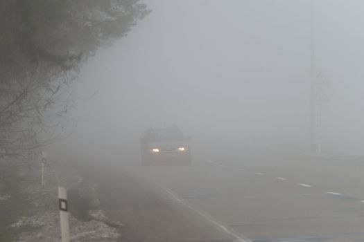 Car traveling on the foggy road with headlights or headlamps on. Low visibility - Dangerous driving of cars in winter in bad weather.