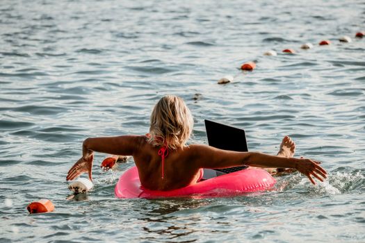 Woman works on laptop in sea. Freelancer, young blond woman in sunglases floating on an inflatable big pink donut with a laptop in the sea at sunset. Freelance, travel and holidays concept