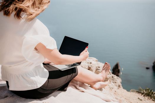 Successful business woman in yellow hat working on laptop by the sea. Pretty lady typing on computer at summer day outdoors. Freelance, travel and holidays concept.