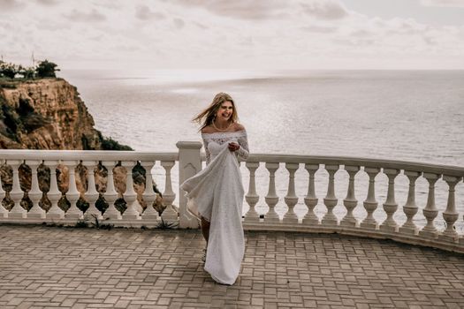 Romantic beautiful bride in white dress posing with sea and mountains in background. Stylish bride standing back on beautiful landscape of sea and mountains on sunset