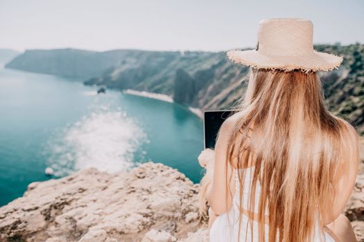 Successful business woman in yellow hat working on laptop by the sea. Pretty lady typing on computer at summer day outdoors. Freelance, travel and holidays concept.