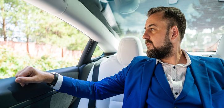 A caucasian man in a blue suit looks out the open window while sitting in the back seat of a car. Business class passenger