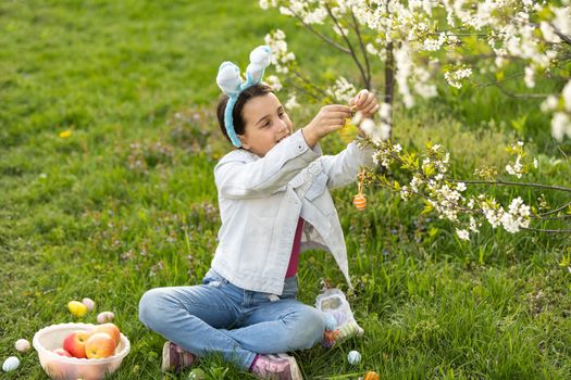 Adorable little girl in bunny ears, blooming tree branch outdoors on a spring day. Kid having fun on Easter egg hunt in the garden.