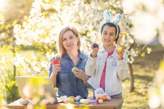 Mother and her daughter painting eggs. Happy family preparing for Easter. Cute little child girl wearing bunny ears.