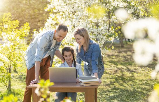Happy family of four parents and cute little girl studying on laptop, enjoy using laptop watching cartoons, make internet video call or shopping online looking at computer screen sit together outdoor.