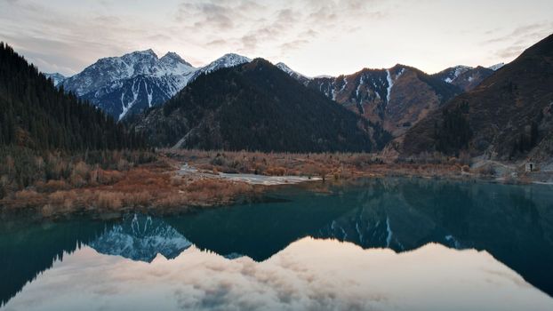 Issyk mountain lake with mirror water at sunset. The color of the water changes before our eyes. There are trees in clear water. Snowy mountains and green hills are visible. Clouds are reflected