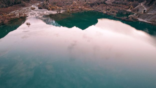 Turquoise blue mirror water with trees in the lake. Light streaks from underwater streams are visible. Autumn mountains and coniferous trees are reflected in the water. Issyk Mountain Lake, Kazakhstan