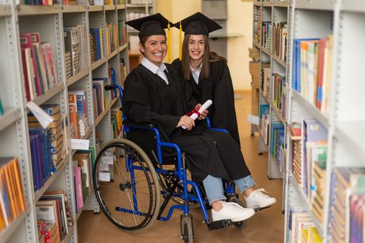 Happy young woman and woman in wheelchair in graduate gown with diploma in hands in library. Inclusive education