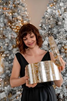 Woman Christmas tree gift. Happy romantic brunette girl in black dress standing on the background of the Christmas tree. Cheerful lady was surprised by the gift after opening the gift box. Merry Christmas and Happy Holidays.