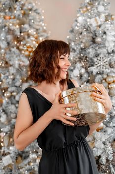 Woman Christmas tree gift. Happy romantic brunette girl in black dress standing on the background of the Christmas tree. Cheerful lady was surprised by the gift after opening the gift box. Merry Christmas and Happy Holidays.
