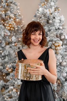 Woman Christmas tree gift. Happy romantic brunette girl in black dress standing on the background of the Christmas tree. Cheerful lady was surprised by the gift after opening the gift box. Merry Christmas and Happy Holidays.