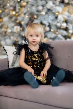 A little girl is sitting on the background of a Christmas tree. Blonde 2 years old in a black dress sits on a beige sofa in a room decorated for christmas and new year. Photo under the tree