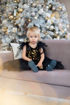 A little girl is sitting on the background of a Christmas tree. Blonde 2 years old in a black dress sits on a beige sofa in a room decorated for christmas and new year. Photo under the tree