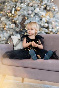 A little girl is sitting on the background of a Christmas tree. Blonde 2 years old in a black dress sits on a beige sofa in a room decorated for christmas and new year. Photo under the tree