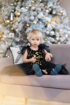 A little girl is sitting on the background of a Christmas tree. Blonde 2 years old in a black dress sits on a beige sofa in a room decorated for christmas and new year. Photo under the tree