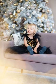 A little girl is sitting on the background of a Christmas tree. Blonde 2 years old in a black dress sits on a beige sofa in a room decorated for christmas and new year. Photo under the tree