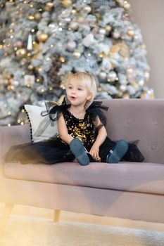 A little girl is sitting on the background of a Christmas tree. Blonde 2 years old in a black dress sits on a beige sofa in a room decorated for christmas and new year. Photo under the tree