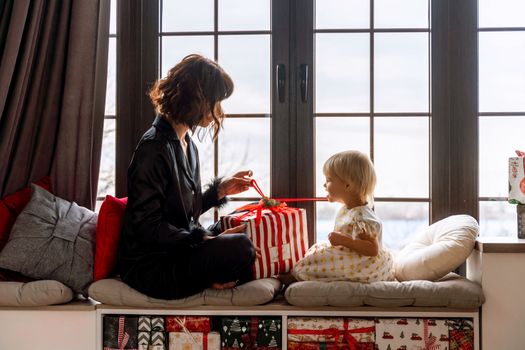 Mother and daughter sit on the windowsill and hold a gift between them. In a decorated Christmas room, mother in black pajamas, daughter in white dress. New Year's family holidays
