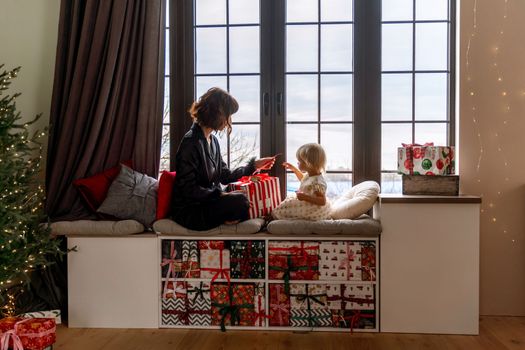 Mother and daughter sit on the windowsill and hold a gift between them. In a decorated Christmas room. New Year's family holidays