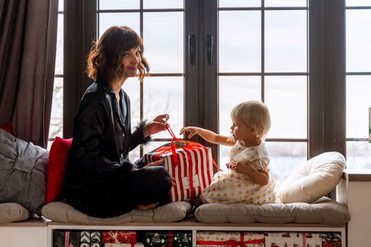 Mother and daughter sit on the windowsill and hold a gift between them. In a decorated Christmas room, mother in black pajamas, daughter in white dress. New Year's family holidays