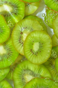 Close-up view of the green kiwi fruit slices in water background. Texture of cooling fruit drink with macro bubbles on the glass wall. Flat design. Vertical image.